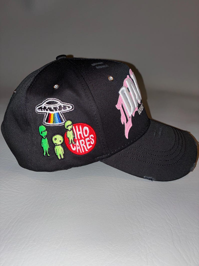 May include: A black baseball cap featuring distressed detailing on the brim and crown. The side displays embroidered patches of a UFO with a rainbow beam, three green alien figures, and a red circular patch with the white text WHO CARES.