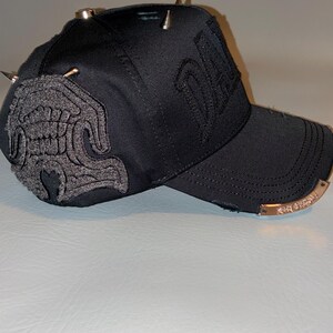 May include: A black baseball cap featuring a textured grey embroidered patch on the side, metallic silver spikes on the crown, and a curved brim with a metallic plate attached to the edge. The cap includes distressed fabric details and embroidery.