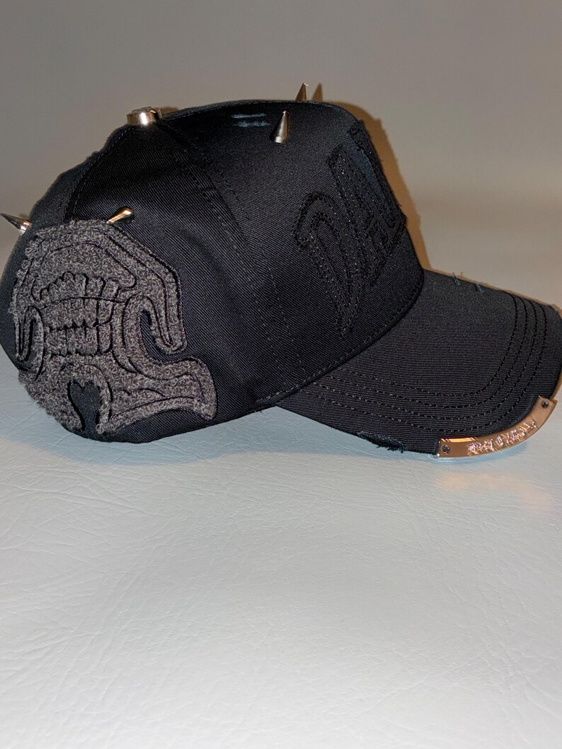 May include: A black baseball cap featuring a textured grey embroidered patch on the side, metallic silver spikes on the crown, and a curved brim with a metallic plate attached to the edge. The cap includes distressed fabric details and embroidery.