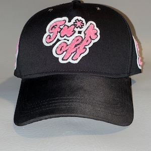Puede incluir: Una gorra de b&eacute;isbol negra con un parche de chenilla rosa y blanco en el panel frontal con el texto "Fn*ck off" en una fuente estilizada. La gorra tiene una corona estructurada, una visera curva y detalles laterales texturizados en rosa.