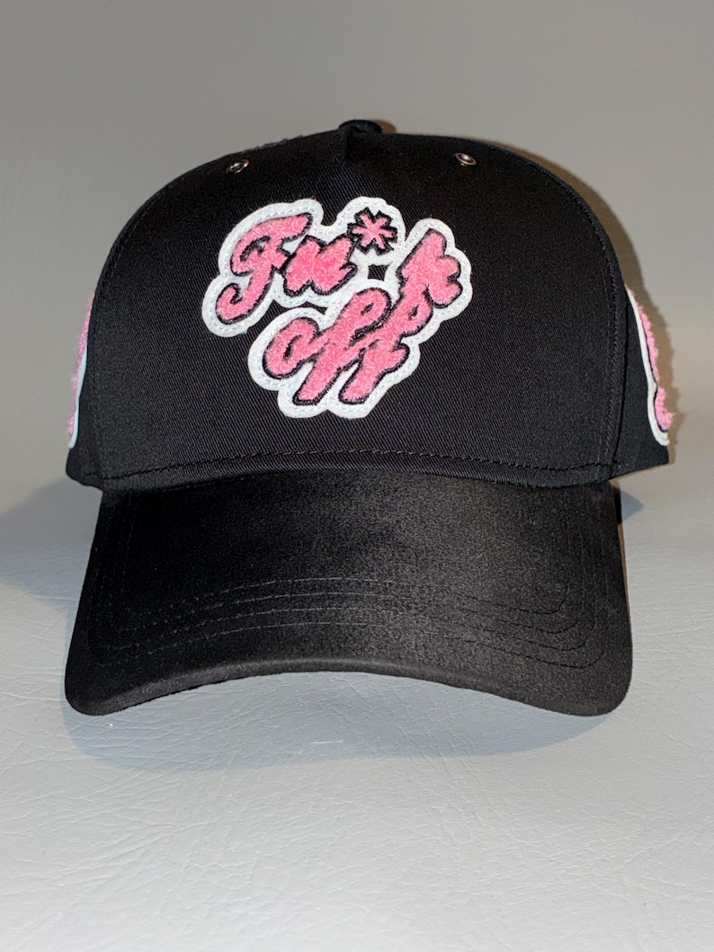 Puede incluir: Una gorra de b&eacute;isbol negra con un parche de chenilla rosa y blanco en el panel frontal con el texto "Fn*ck off" en una fuente estilizada. La gorra tiene una corona estructurada, una visera curva y detalles laterales texturizados en rosa.