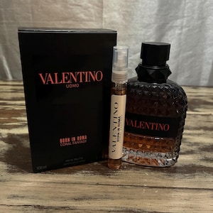 Op de afbeelding: Een zwarte doos en een donkerbruine parfumfles met de tekst "VALENTINO UOMO BORN IN ROMA CORAL FANTASY" in het rood. Een kleine parfummonster is ook inbegrepen. De parfumfles heeft een zwarte dop en een textuurontwerp.
