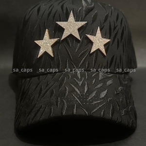May include: Black baseball cap with a textured leaf pattern. The cap features three gold-colored star embellishments encrusted with rhinestones. The brim is curved, and the cap has a classic six-panel design.