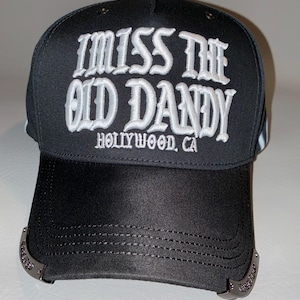 Puede incluir: Una gorra de b&eacute;isbol negra con texto bordado en blanco que dice I MISS THE OLD DANDY HOLLYWOOD, CA. La gorra tiene una corona estructurada y una visera curva con detalles decorativos en tono plateado met&aacute;lico en los bordes laterales.