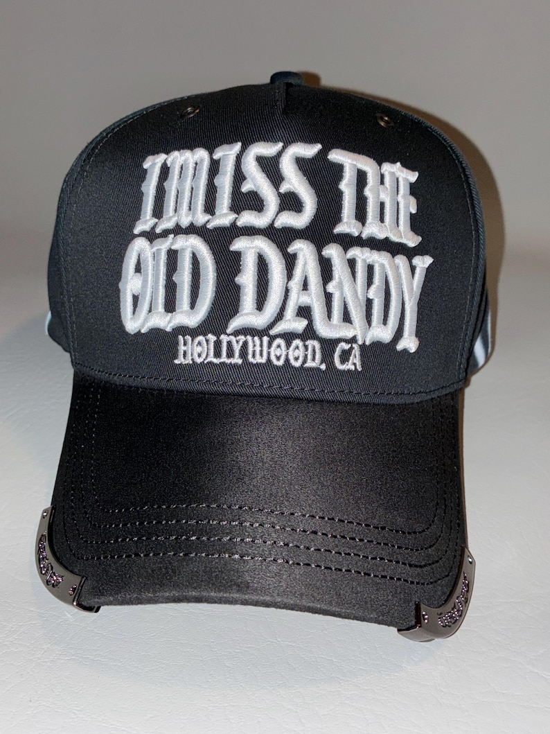 Puede incluir: Una gorra de b&eacute;isbol negra con texto bordado en blanco que dice I MISS THE OLD DANDY HOLLYWOOD, CA. La gorra tiene una corona estructurada y una visera curva con detalles decorativos en tono plateado met&aacute;lico en los bordes laterales.