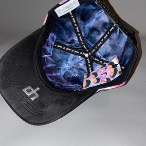 Puede incluir: Una gorra de b&eacute;isbol negra con un logotipo plateado en la visera. El interior presenta un forro con patr&oacute;n de galaxia azul y morado con texto repetido. Una etiqueta blanca dice BAD BOYZ BY DANDYHATS, y una etiqueta colgante muestra caras de dibujos animados y el texto BAD BOYZ.