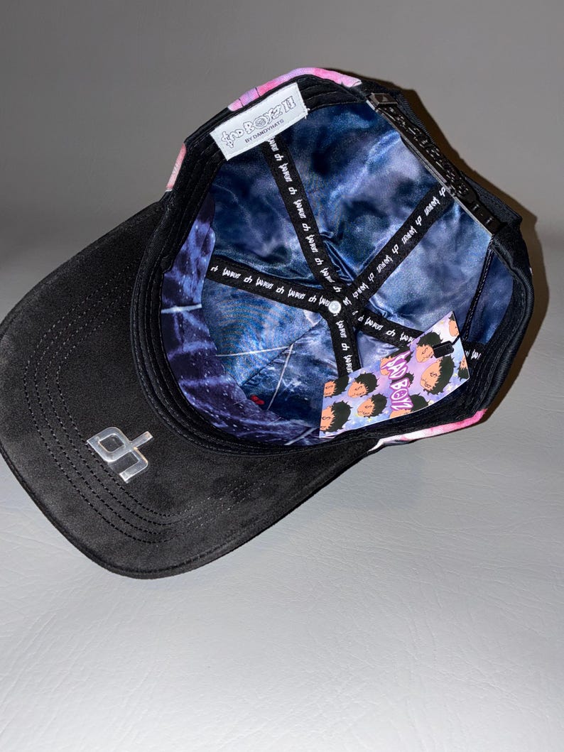 Puede incluir: Una gorra de b&eacute;isbol negra con un logotipo plateado en la visera. El interior presenta un forro con patr&oacute;n de galaxia azul y morado con texto repetido. Una etiqueta blanca dice BAD BOYZ BY DANDYHATS, y una etiqueta colgante muestra caras de dibujos animados y el texto BAD BOYZ.