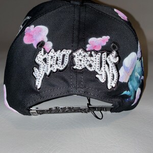 Puede incluir: Una gorra de b&eacute;isbol negra con el texto SAD BOYS en letras blancas con tachuelas en la parte delantera. La gorra est&aacute; decorada con un estampado de nubes rosas y moradas y criaturas azuladas. Incluye una correa trasera negra ajustable.