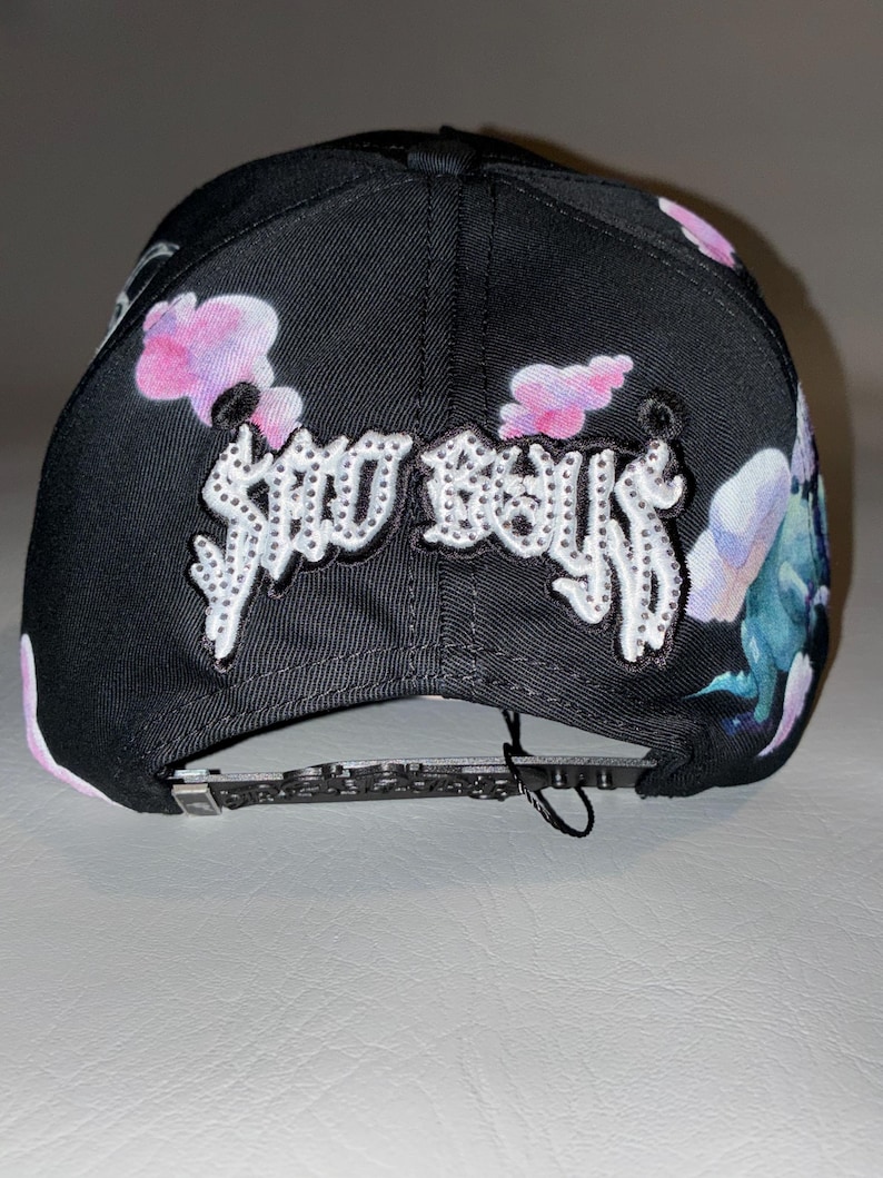 Puede incluir: Una gorra de b&eacute;isbol negra con el texto SAD BOYS en letras blancas con tachuelas en la parte delantera. La gorra est&aacute; decorada con un estampado de nubes rosas y moradas y criaturas azuladas. Incluye una correa trasera negra ajustable.