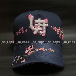 May include: Navy blue baseball cap with embroidered Japanese characters and cherry blossom accents. The cap features the text "LOS ANGELES" in pink and the brand name "_sa_caps" repeated along the brim.