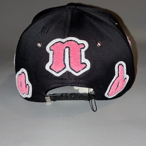 Puede incluir: Una gorra de b&eacute;isbol negra con una letra n rosa grande y central con borde blanco, flanqueada por letras rosas m&aacute;s peque&ntilde;as a los lados. La gorra incluye ojales met&aacute;licos de color plateado y un cierre trasero ajustable de color negro.