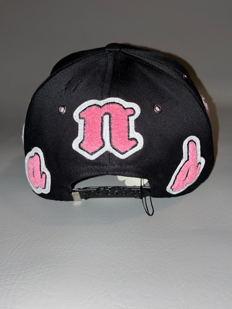 Puede incluir: Una gorra de b&eacute;isbol negra con una letra n rosa grande y central con borde blanco, flanqueada por letras rosas m&aacute;s peque&ntilde;as a los lados. La gorra incluye ojales met&aacute;licos de color plateado y un cierre trasero ajustable de color negro.