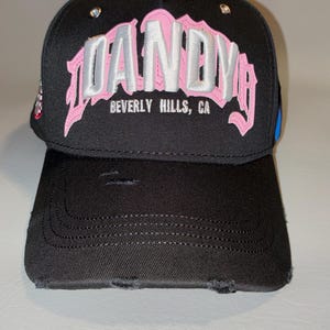 May include: A black baseball cap with a distressed, worn-in finish. The front features large, embroidered lettering in pink and silver that reads DANDY, with the text BEVERLY HILLS, CA printed in smaller white letters underneath the main design.