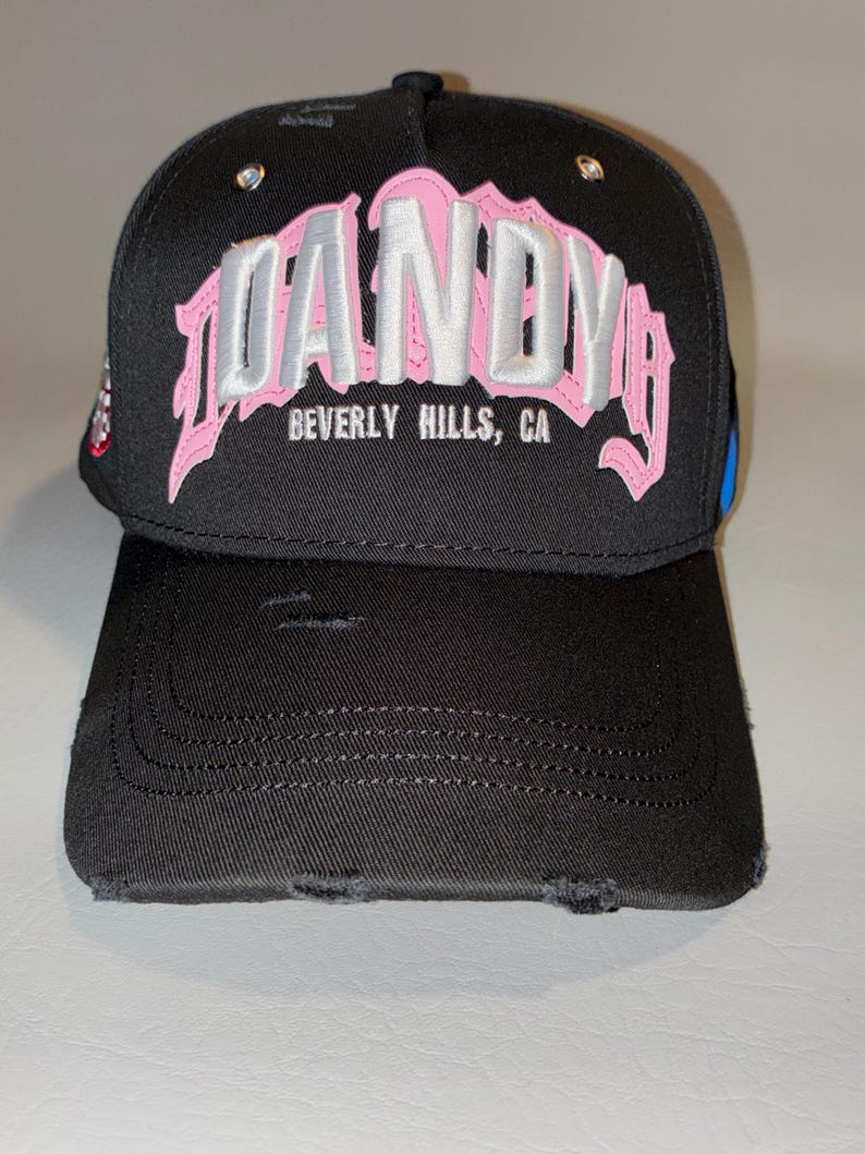 May include: A black baseball cap with a distressed, worn-in finish. The front features large, embroidered lettering in pink and silver that reads DANDY, with the text BEVERLY HILLS, CA printed in smaller white letters underneath the main design.