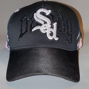 Puede incluir: Una gorra de b&eacute;isbol negra con la palabra "SAD" en letras grandes de color blanco incrustadas con pedrer&iacute;a en el panel frontal. Los laterales de la gorra muestran un estampado floral colorido y la visera es de un material negro liso tipo ante.