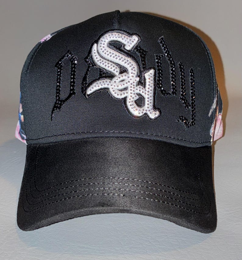 Puede incluir: Una gorra de b&eacute;isbol negra con la palabra "SAD" en letras grandes de color blanco incrustadas con pedrer&iacute;a en el panel frontal. Los laterales de la gorra muestran un estampado floral colorido y la visera es de un material negro liso tipo ante.