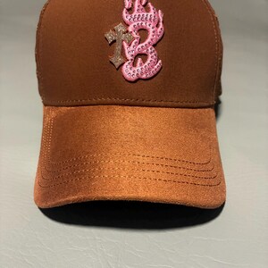 May include: A brown baseball cap featuring a textured suede-like brim and a smooth crown. The front displays an embroidered pink flame-shaped letter B embellished with small rhinestones, positioned next to a glittery silver-toned cross emblem.