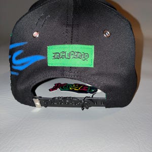 May include: A black baseball cap featuring a bright green rectangular patch with the text DANDY in a gothic font. The cap includes blue flame-like graphics on the side, silver metal eyelets, and a black plastic snapback closure with embossed text.