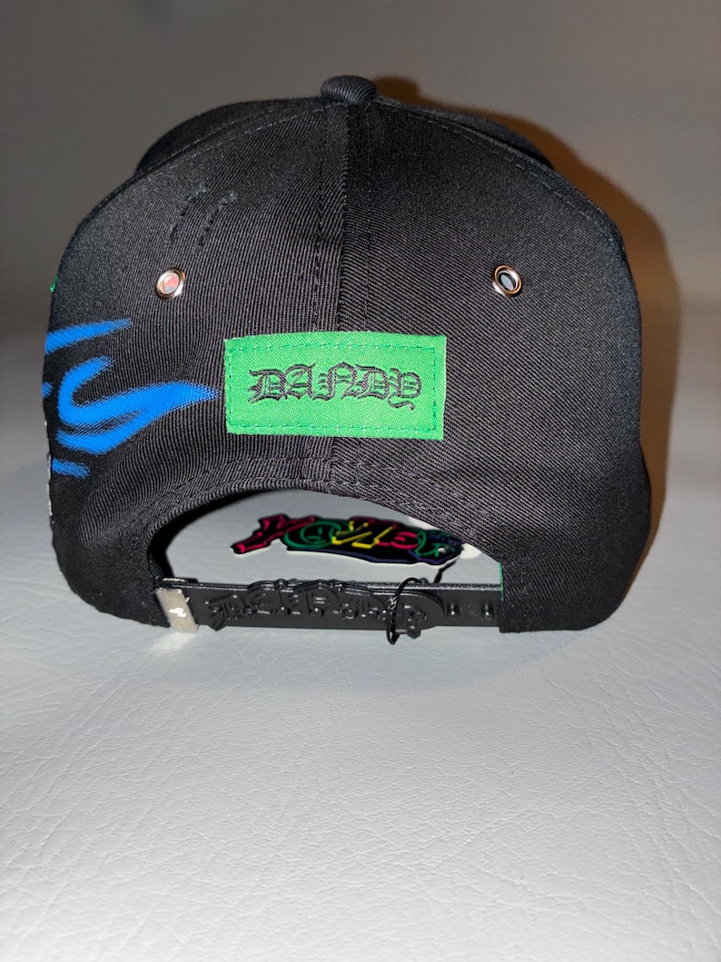 May include: A black baseball cap featuring a bright green rectangular patch with the text DANDY in a gothic font. The cap includes blue flame-like graphics on the side, silver metal eyelets, and a black plastic snapback closure with embossed text.