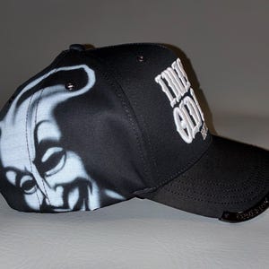 Puede incluir: Una gorra de b&eacute;isbol negra con un gr&aacute;fico en blanco aerografiado de una cara sonriente en el panel lateral. La parte frontal muestra el texto bordado en blanco "LUCKY ODD" y un peque&ntilde;o detalle met&aacute;lico en el borde de la visera.