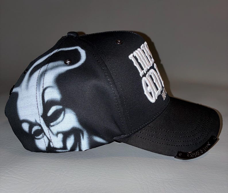 Puede incluir: Una gorra de b&eacute;isbol negra con un gr&aacute;fico en blanco aerografiado de una cara sonriente en el panel lateral. La parte frontal muestra el texto bordado en blanco "LUCKY ODD" y un peque&ntilde;o detalle met&aacute;lico en el borde de la visera.