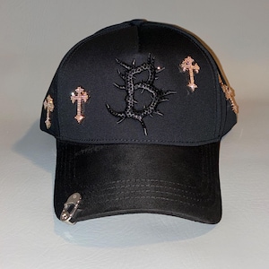 May include: A black baseball cap featuring a central letter B with a thorny texture, surrounded by four metallic cross emblems with embedded crystals. A silver-toned metal ring is attached to the left side of the curved brim of the cap.