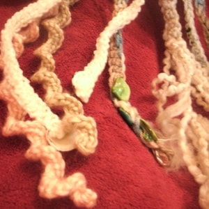 May include: A close-up of a multi-colored crocheted cord with a green and blue bead. The cord is made of different textures and colors, including white, pink, and brown.