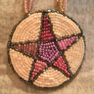 May include: A beaded pendant with a five-pointed star design. The star is made of pink, purple, and red beads, and is outlined in black beads. The pendant is surrounded by white beads.