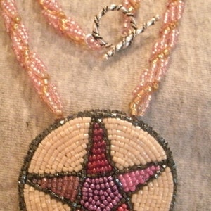 May include: A beaded necklace with a pink and gold chain and a round pendant featuring a five-pointed star in shades of pink, red, and gray.