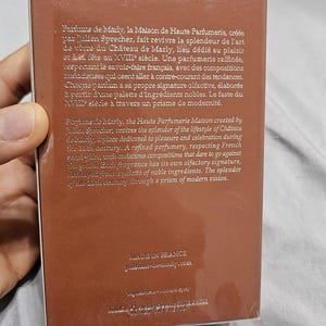 May include: A brown rectangular perfume box with text in English and French. The box is held by a person's hand. The text describes the brand's history and fragrance notes.