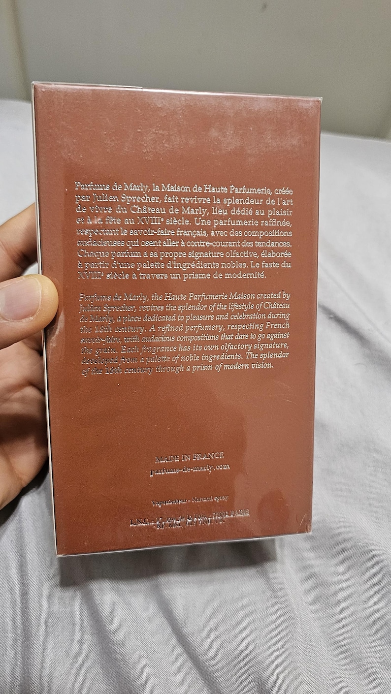 May include: A brown rectangular perfume box with text in English and French. The box is held by a person's hand. The text describes the brand's history and fragrance notes.
