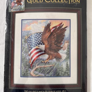 May include: A Dimensions Gold Collection counted cross stitch kit titled "Flight of Freedom." The design features an American bald eagle with outstretched wings, the American flag, and a mountain landscape. The finished size is 12 x 14 inches.