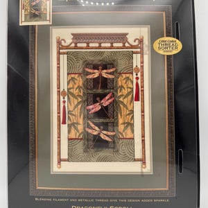 May include: A Dimensions Gold Collection counted cross-stitch kit titled "Dragonfly Scroll." The design features three dragonfly images in shades of pink and brown, set against a green and beige background. The kit measures 11" x 16" (28 x 42 cm).