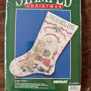 May include: A cross-stitch kit for a Christmas stocking featuring a Santa Claus with two teddy bears. The stocking is white with red and green accents. The kit includes stamped fabric, floss, needles, and instructions. The kit is titled "Talking to Santa" and is 9 inches by 16 inches. The kit is made by Bernat Crafts.