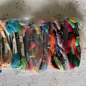 May include: A collection of embroidery floss in various colors, including orange, teal, pink, and green, packaged in clear plastic bags. The floss is labeled with product information.
