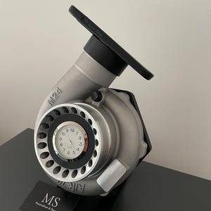 Puede incluir: Un turbocompresor plateado reutilizado como reloj y soporte para teléfono. La esfera del reloj es blanca con números negros. El teléfono descansa en la parte superior. La base tiene una tarjeta de visita con el texto "MS Konstruktion & Design".