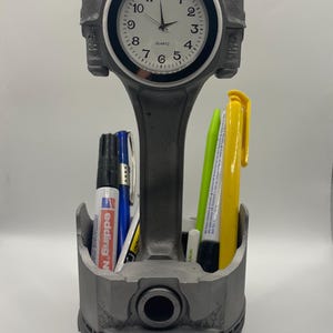 Könnte beinhalten: Ein einzigartiger Schreibtisch-Organizer aus einem Motor-Kolben und einer Pleuelstange gefertigt. Die Oberseite zeigt eine Uhr mit weißem Zifferblatt und schwarzen Ziffern. Die Basis enthält Stifte und Marker, darunter ein blauer und roter Edding N Permanentmarker. Ein kreatives und funktionales Schreibtischzubehör.
