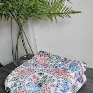 May include: Folded white fabric with a colourful floral pattern in red, blue, and green. The fabric is embroidered with a detailed design. A vase of green fern-like plants is in the background.