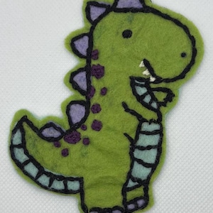 Könnte beinhalten: Ein grünes Dinosaurier-Applikation aus Filz mit lila Akzenten. Der Dinosaurier hat eine schwarze Umrandung, lila Stacheln und lila Punkte. Bauch und Schwanz haben blau-schwarze Streifen. Auf weißem Hintergrund.