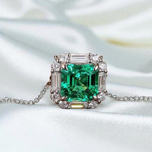 May include: A pendant necklace featuring a square-cut emerald gemstone, encircled by a halo of diamonds. The emerald is a vivid green, set within a frame of smaller, clear diamonds. The chain is a delicate silver.
