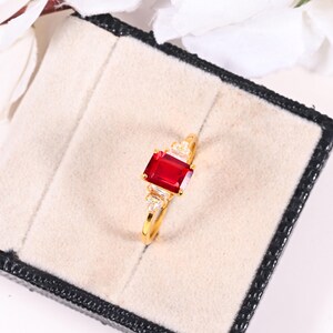 May include: A gold ring with a large, rectangular, deep red gemstone at its centre. The ring is embellished with smaller, clear gemstones on either side of the main stone, set against a neutral background.