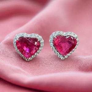 May include: A pair of heart-shaped earrings with a vibrant pink gemstone center, surrounded by a halo of small, sparkling clear stones. The earrings rest on a soft, pink fabric, creating a romantic and elegant display.