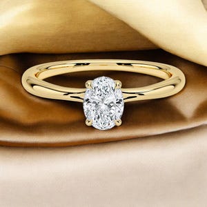 以下が含まれることがあります： 大きな楕円形のダイヤモンドが中央にセットされたゴールドリング。 バンドは滑らかで磨かれたゴールドです。 ダイヤモンドは透明で輝き、4つの爪で留められています。 リングは金と茶色の生地の上に展示されています。