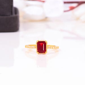 以下が含まれることがあります： 中央に長方形の深い赤色の宝石をあしらったゴールドリング。バンドもゴールドで、小さな透明な石が側面にセットされています。リングは白い背景に配置されています。