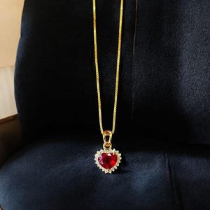 Puede incluir: Un collar dorado con un colgante en forma de corazón. El colgante presenta una gema roja vibrante rodeada de pequeñas piedras transparentes. El collar cuelga sobre un fondo azul oscuro.
