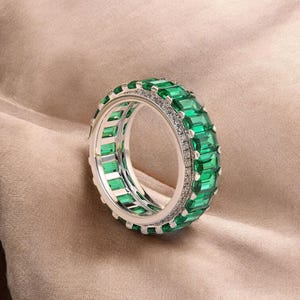 以下が含まれることがあります： エメラルドカットの緑色の宝石が連続したバンドを特徴とするシルバーリング。リングは、緑色の宝石の間に小さなクリアストーンの列があるダブルバンドデザインです。リングはニュートラルな背景にセットされています。