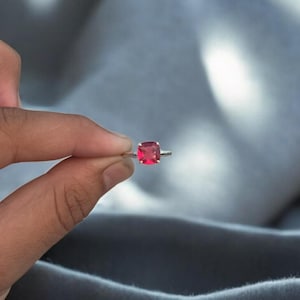 Op de afbeelding: Een zilveren ring met een vierkante, helderrode edelsteen. De edelsteen is het middelpunt, gezet op een dunne, gepolijste band. De ring wordt getoond tegen een zachte, grijze achtergrond.