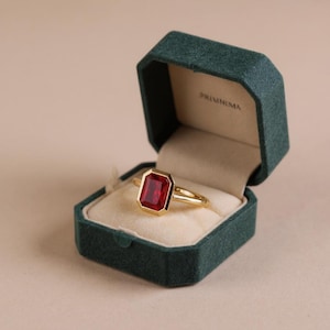 May include: A gold ring with a large, rectangular, red gemstone, set in a gold bezel. The ring is displayed in an open, dark green velvet ring box. The box has the word "PRISINIMA" printed on the inside.