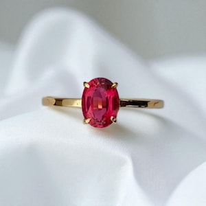 May include: A gold ring with a vivid, oval-shaped red gemstone. The gemstone is secured by four small prongs. The ring's band is a slender, polished gold. The ring is displayed on a white, textured surface.