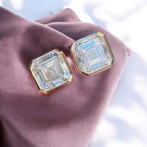 Op de afbeelding: Een paar vierkant geslepen, heldere edelsteen oorbellen met goudkleurige randen. De oorbellen worden gepresenteerd op een mauve satijnen stof, wat een luxueuze en elegante presentatie creëert. De edelstenen hebben een veelzijdig ontwerp.